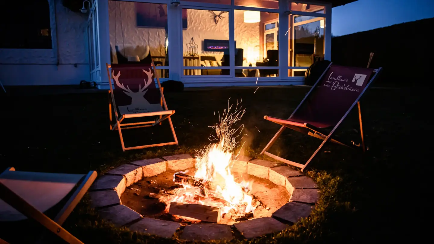 Startseite von landhaus-buechelstein.de: Abend im Garten des Ferienhauses mit Feuerstelle und Liegestühlen, dahinter der beleuchtete Wintergarten
